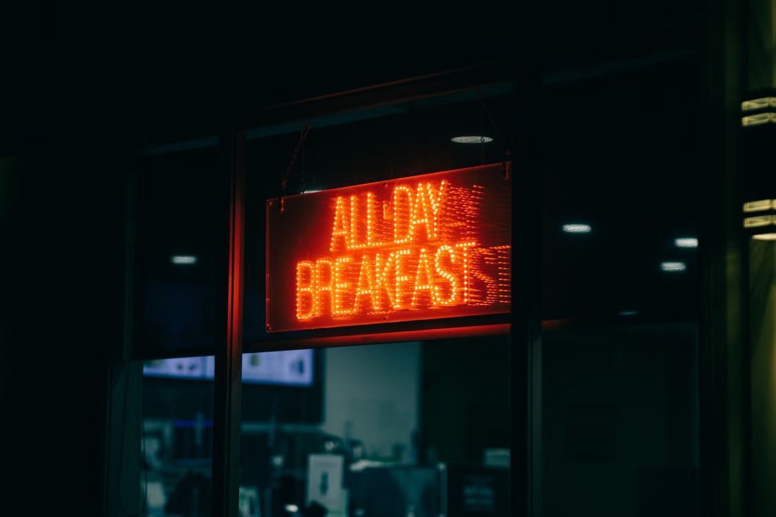 A neon all day breakfast sign glowing at night, welcoming casual date.