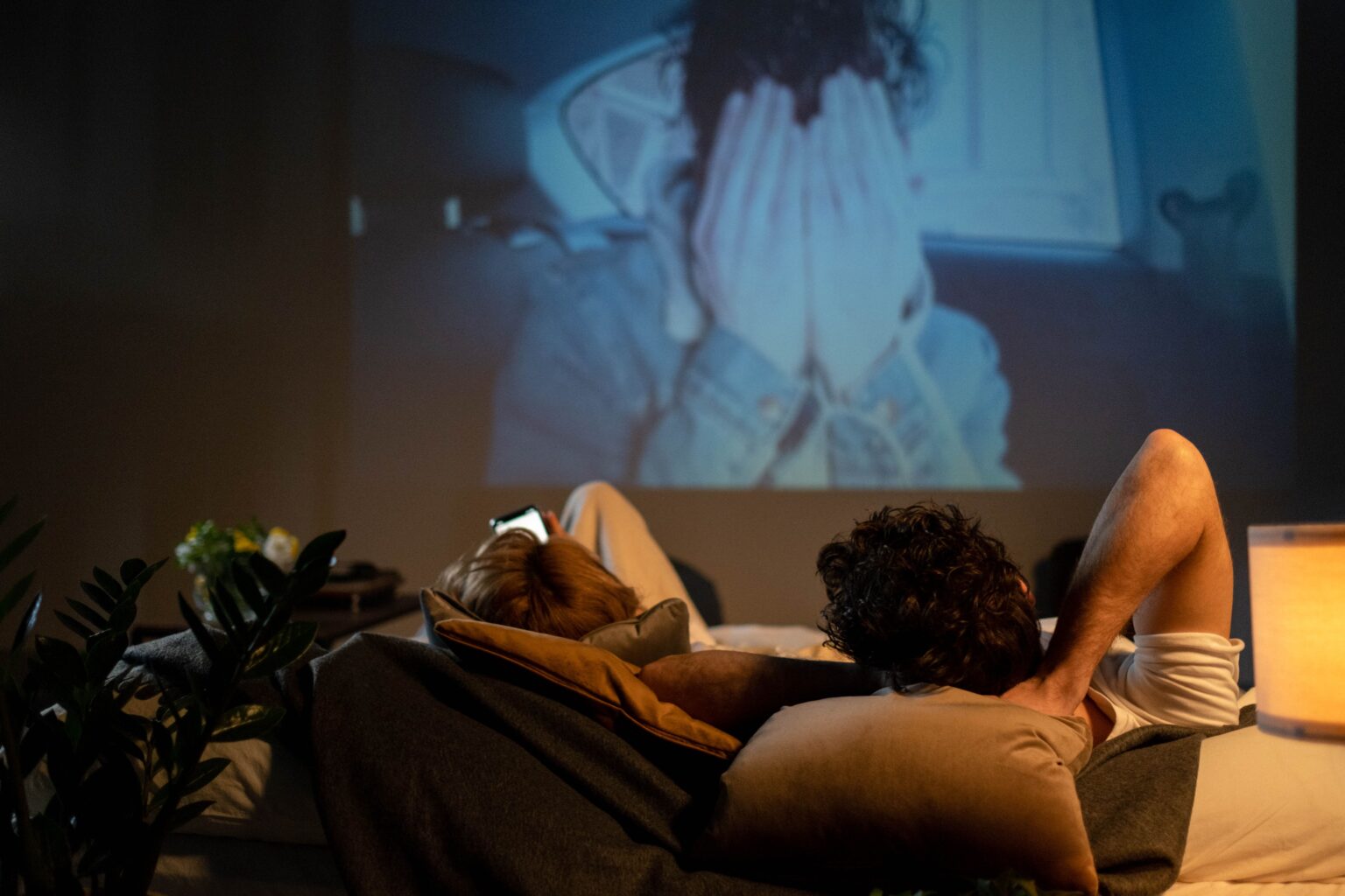 Here Are Date Ideas That We’re Officially Banning “Idk, What Do You Want to Do?” A couple lying in bed watching a movie projector, moving away from boring routine date ideas.