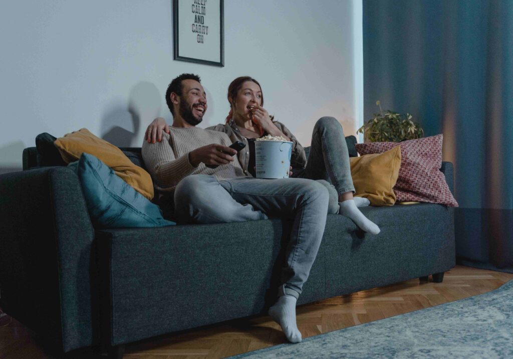A couple laughing on a couch with popcorn, enjoying a movie for fun cozy evening date activities.