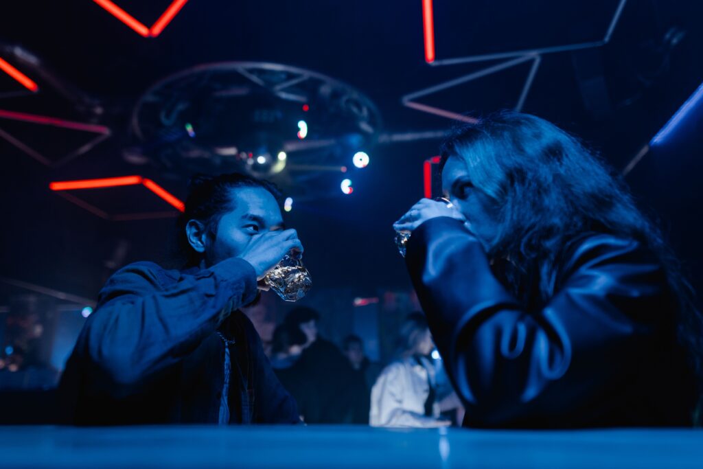 A man and a woman drinking shots at a neon lit bar counter, exchanging funny dark pick up phases.