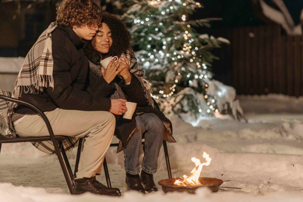 A couple wrapped in a blanket by a fire pit, showcasing cozy outdoor romantic night plans in the snow.