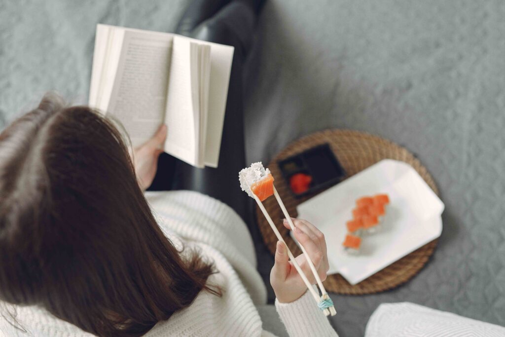 Top view of a woman reading a book and eating sushi on a grey couch, showing peace after no contact with parents.