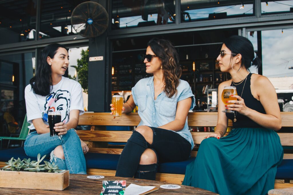 Three female friends talking and holding beers at an outdoor bar bench, showing a supportive circle vs pick me girls.