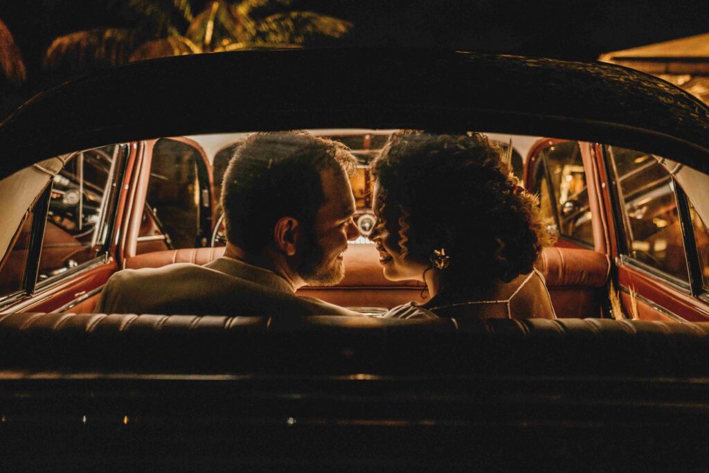 A couple smiling intimately inside a vintage car at night, bonding over their shared twisted sense of humor.