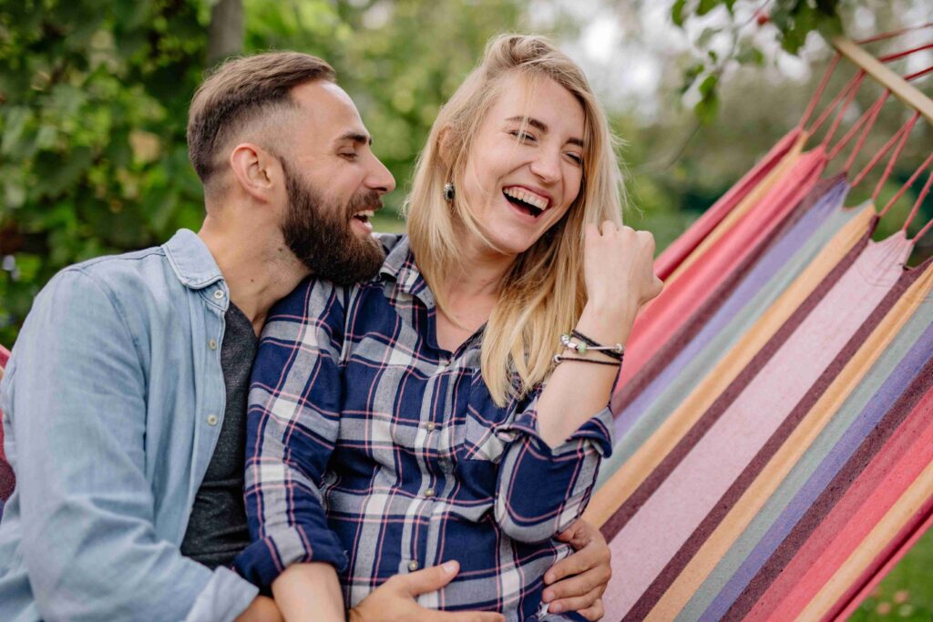 A happy couple laughing together while sitting on a colorful hammock, teasing each other with nsfw pick up lines.